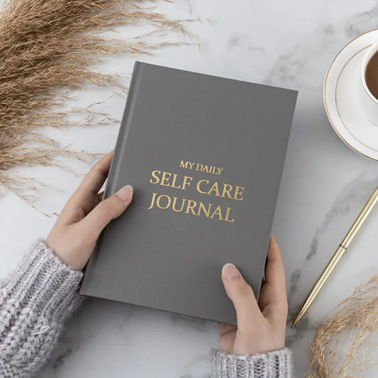 Gray 'My Daily Self Care Journal' held by hands on a marble surface with a cup of coffee and pen, for gratitude and mindfulness. Mental health practice