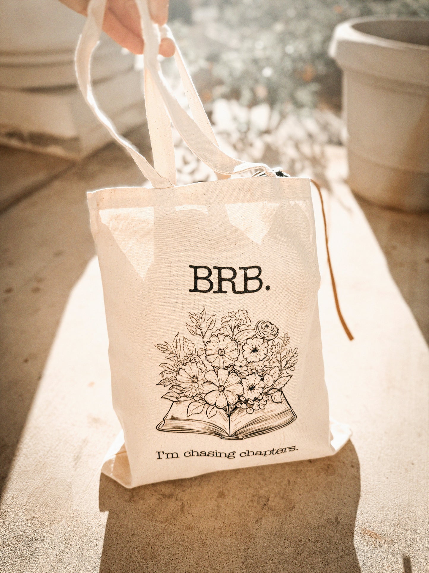 Tote bag with floral design in the center and text that says, "BRB. I'm Chasing Chapters.", held by a hand outdoors