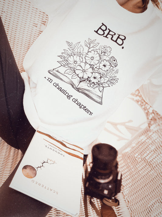 White sweatshirt with text that says "BRB. I'm Chasing Chapters.", with an design of an open book with flowers blooming out of it in the center of the sweatshirt